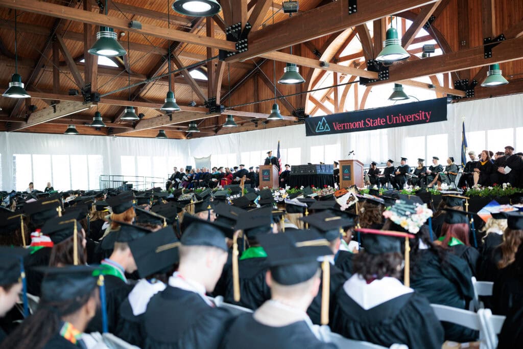 A wide view of the Vermont State University Castleton commencement ceremony inside a large timber-framed pavilion. Hundreds of graduates in black caps and gowns face a stage where a speaker stands at a podium beneath a Vermont State University banner.
