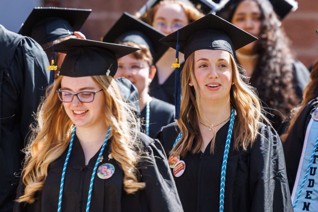 Two smiling female graduates stand close together in black caps and gowns with blue-and-white honor cords. Their caps display gold "2026" tassels. A diverse group of classmates is visible behind them.