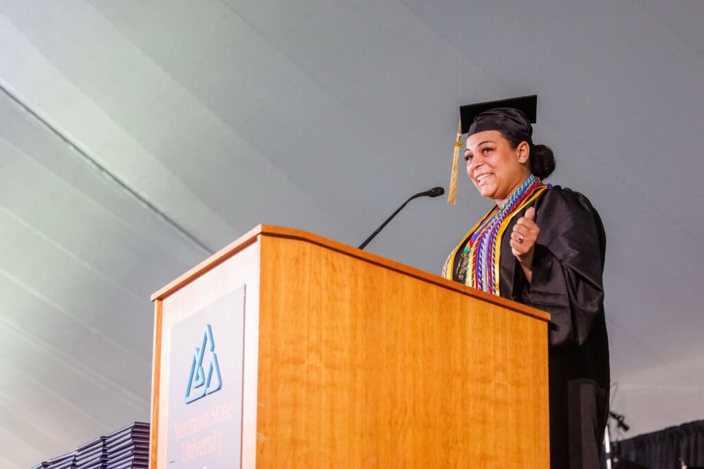 A graduate wearing a black cap and gown adorned with multiple colorful honor cords and medallions delivers a speech at a Vermont State University podium, smiling and gesturing with one hand. The photo is taken from a low angle beneath a tent canopy.