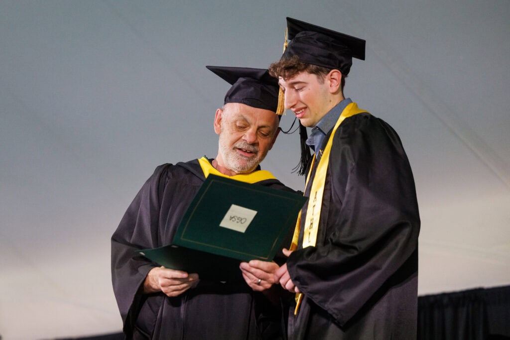 Two people in black caps and gowns with gold accents examine an award together on stage. An older faculty member and a younger graduate lean in to read the document.