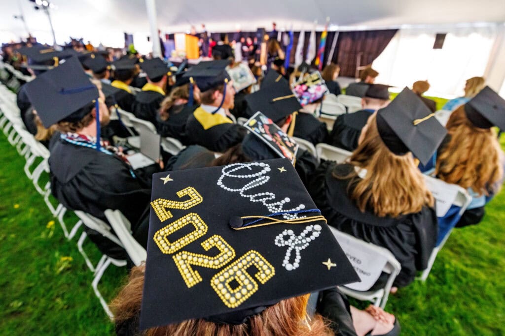 An overhead view of graduates seated at a commencement ceremony under a large tent. In the foreground, a cap is decorated with "2026" in gold rhinestones and a cursive message in white pearls. A colorful stage is visible at the front.