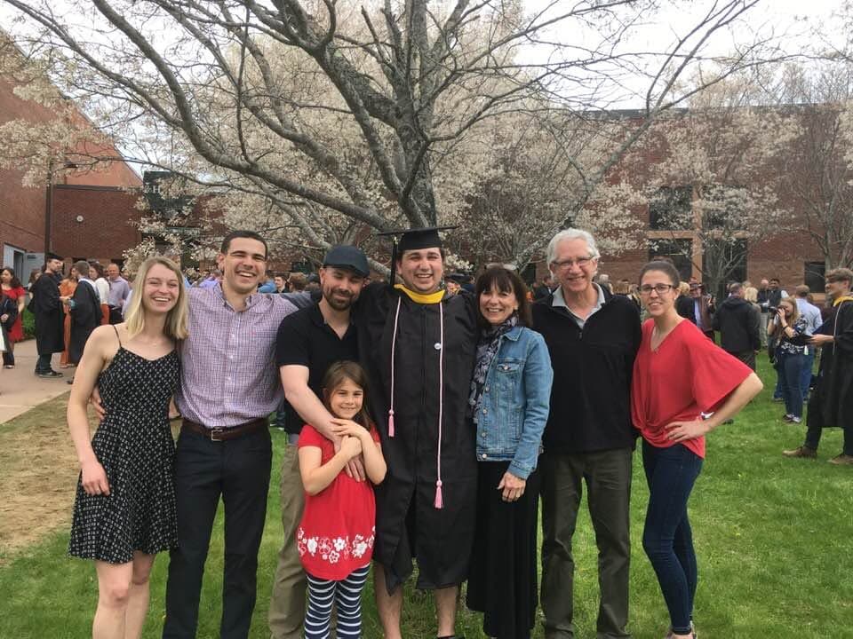 
A group of people stand together outdoors on a college campus during a graduation ceremony. At the center, one individual wears a black graduation cap and gown with a gold stole and tassel, standing as the focal point. Several others stand closely on either side, forming a line, and a young child stands in front near the middle. Behind the group, a tree with white blossoms spreads its branches, and a red-brick academic building is partially visible. In the background, many other attendees and graduates gather on a grassy lawn and walkway. The scene takes place in daylight and suggests a spring commencement event.