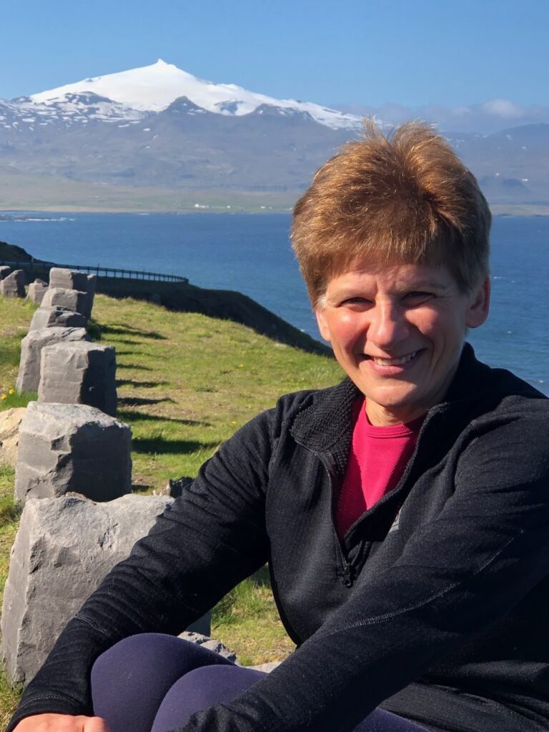 A smiling woman with short auburn hair sits outdoors on ancient stone ruins, wearing a black fleece jacket over a pink shirt. Behind her, a sweeping Icelandic landscape unfolds with a bright blue fjord, green coastal hills, and the snow-capped Snæfellsjökull glacier volcano rising in the distance under a clear blue sky.