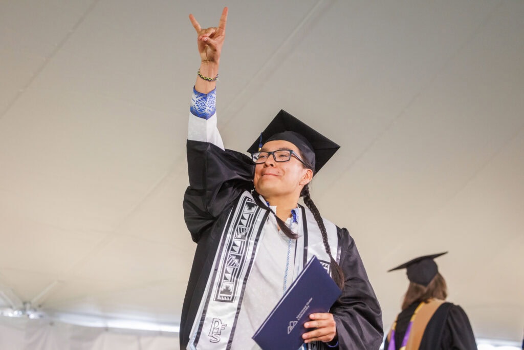 A student in a cap and gown raising their hand above their head in celebration while holding their diploma