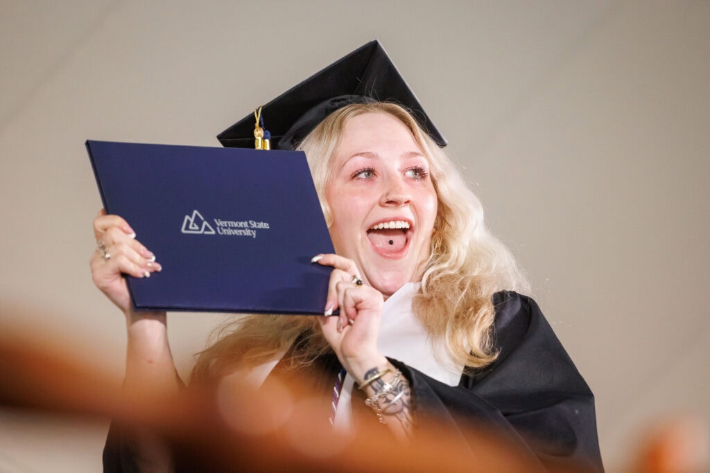 A young woman in graduation cap and gown grinning while holding her diploma 