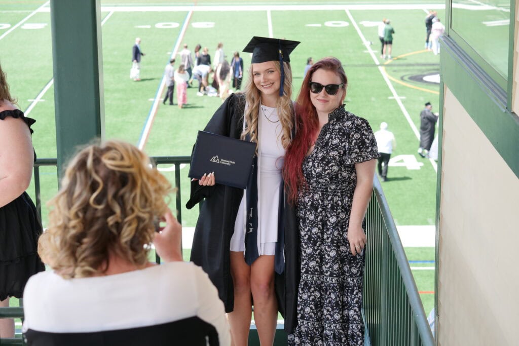 A young woman in a cap and gown holding her diploma while posing with a family member
