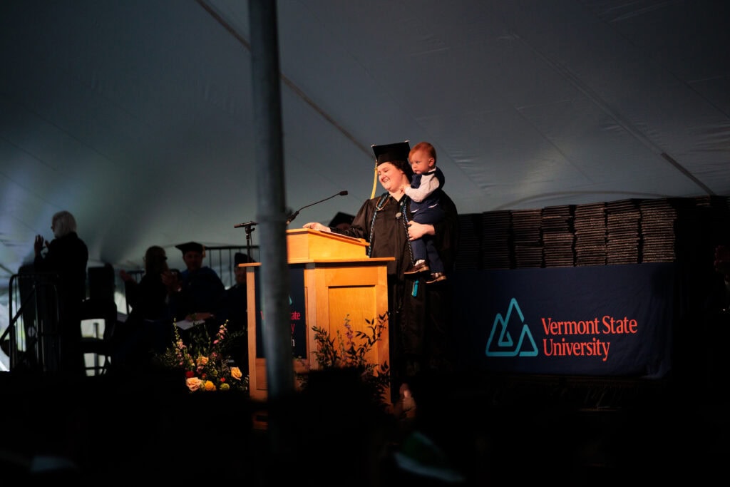 A mother in graduation regalia holding her young child in her arms and standing at a lectern