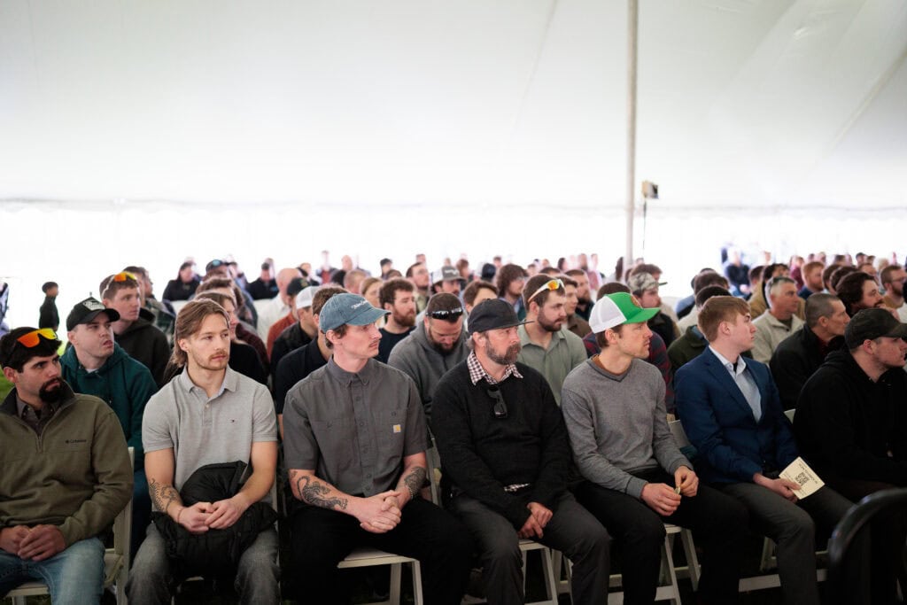 A large crowd of apprentice graduates and guests seated in rows of white folding chairs under a large white tent, attentively facing a stage or speaker out of frame. Many attendees wear baseball caps and casual attire.
