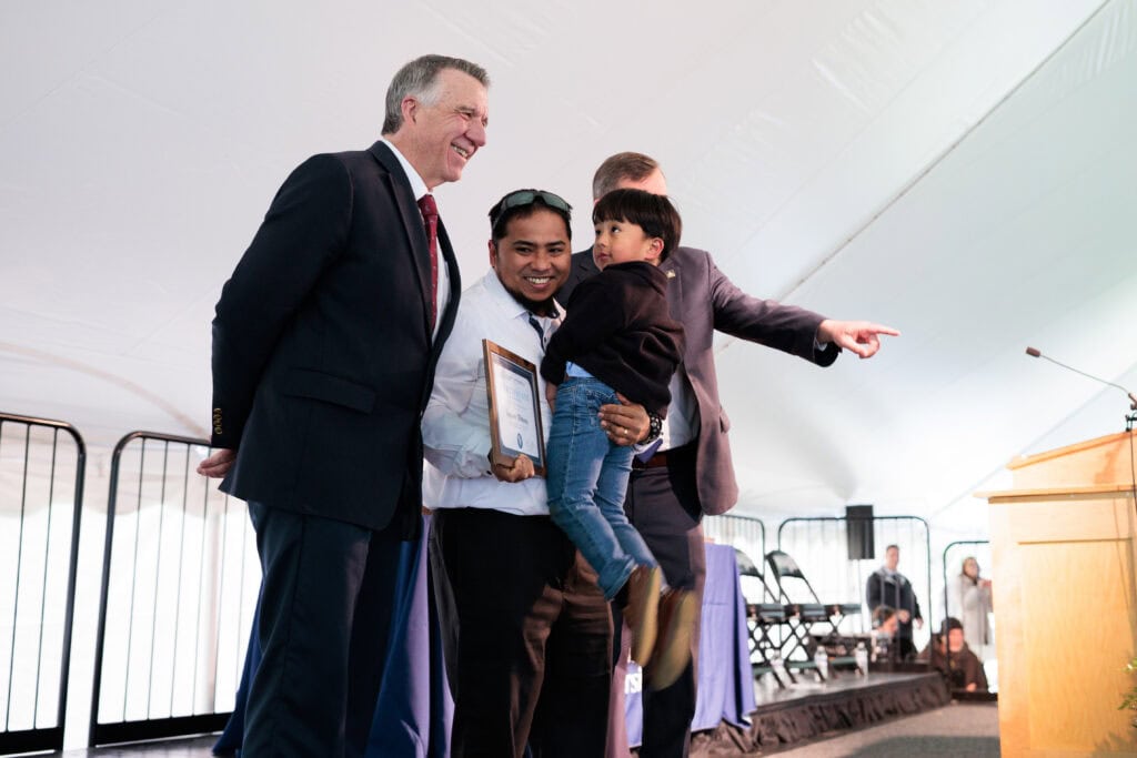 A graduate holds his framed certificate and poses with a woman beside him while family members photograph them outdoors, surrounded by bare trees and other graduates celebrating nearby.