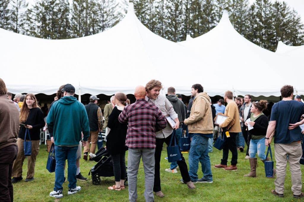 Graduates and guests mingle on a grassy lawn outside a large white tent following the ceremony, many carrying navy blue tote bags and framed certificates, sharing congratulations and conversation.
