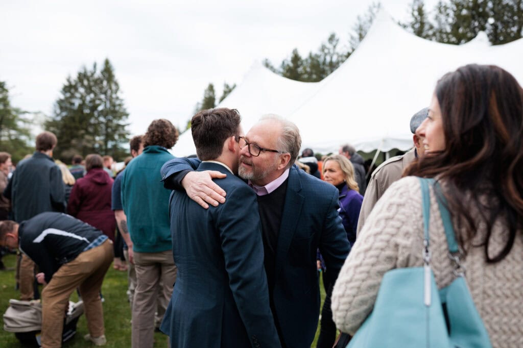 A smiling young man embraces an older man in a warm hug among a seated crowd inside a white tent, as a woman nearby captures the moment on her phone.