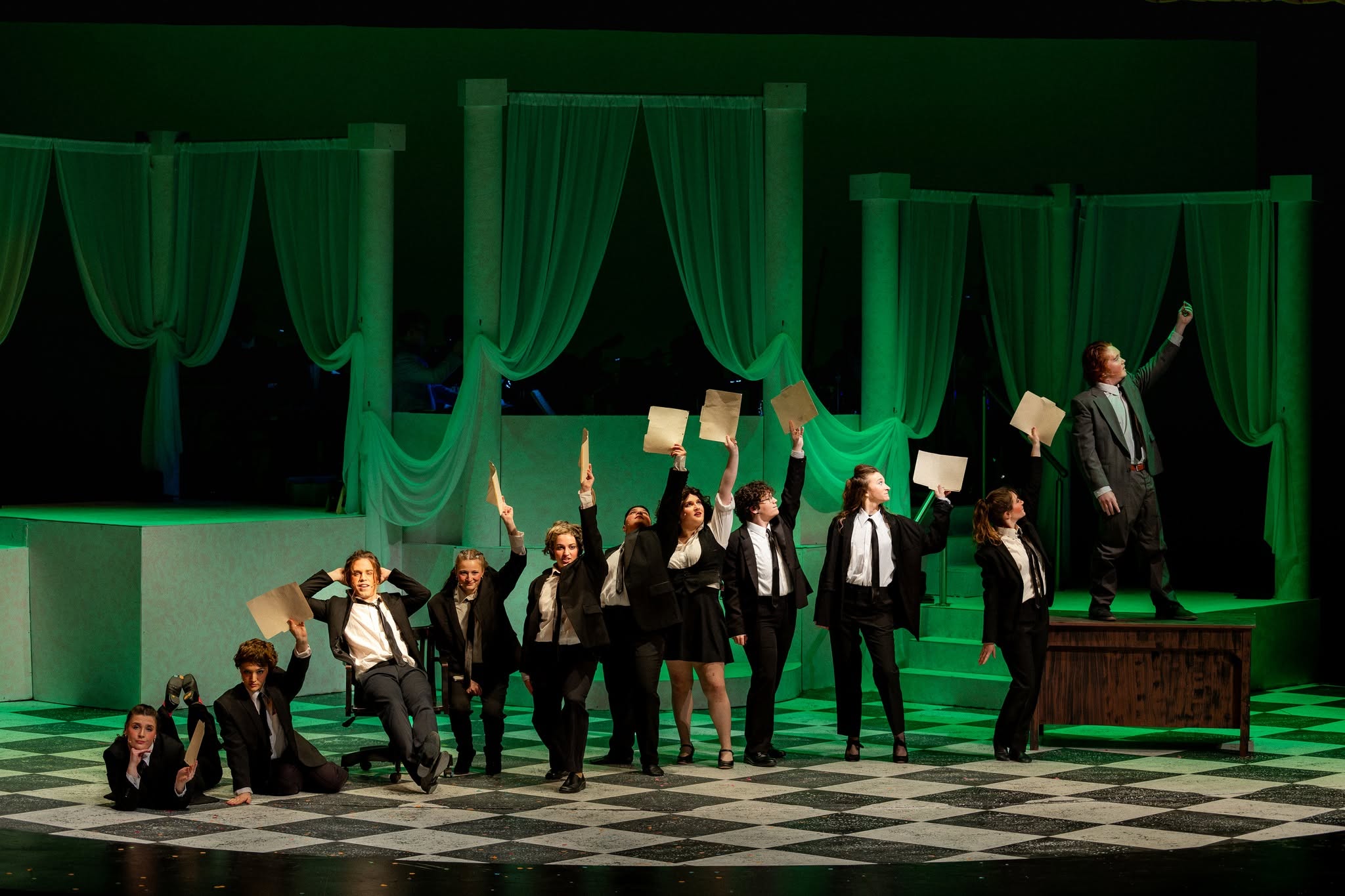 A group of dancers dressed in suits standing in a line representing a bar graph, with a man standing on a desk at the tallest point