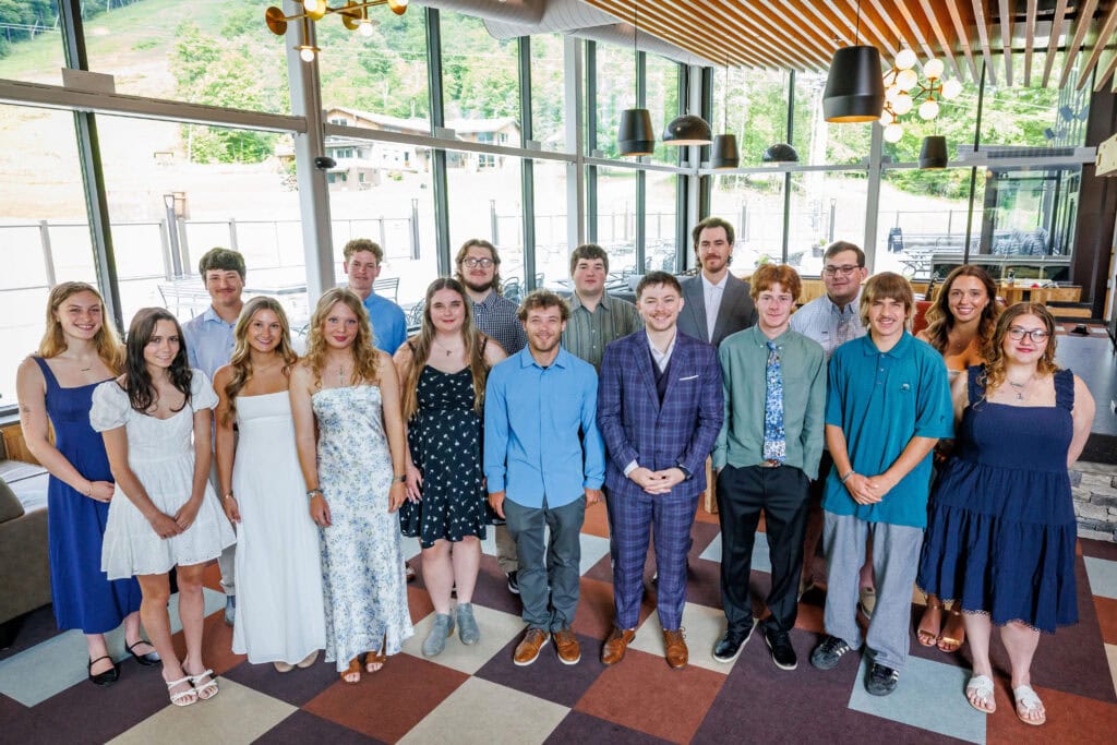Group portrait of about sixteen young graduates standing together indoors, dressed in semi-formal attire — sundresses, button-downs, and one man in a plaid suit. Floor-to-ceiling windows behind them show a grassy ski slope and nearby buildings