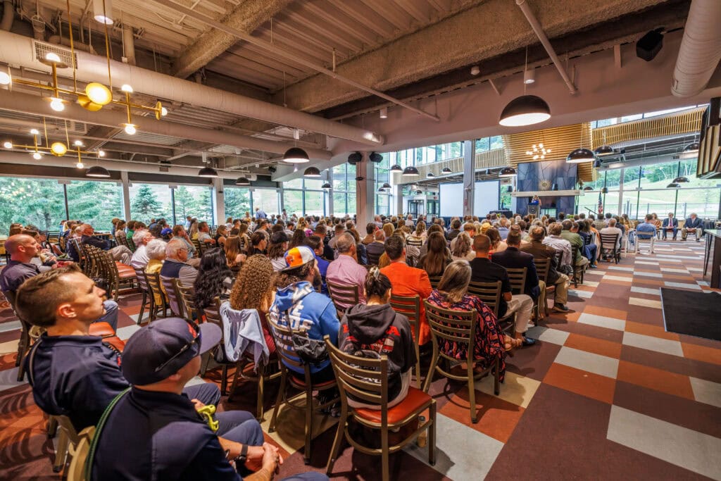 Wide view of a seated audience at an indoor ceremony, viewed from behind, facing a stage area with a stone fireplace and a woman speaking at a podium. Large windows on the left show trees and a ski slope outside.