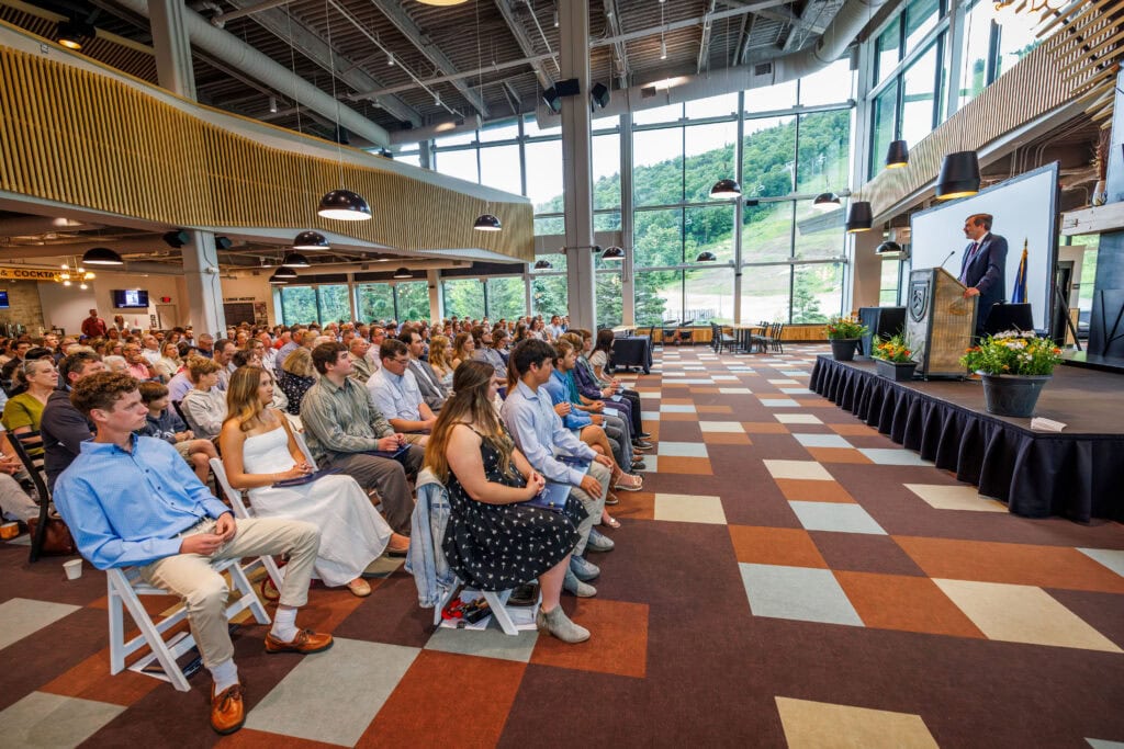 Audience of graduates and family members seated in rows facing a stage, where a man in a suit speaks at a wooden podium displayed on a large screen. Floor-to-ceiling windows behind the stage show a green ski slope and chairlift. Young graduates in the front rows wear light-colored dresses and button-down shirts