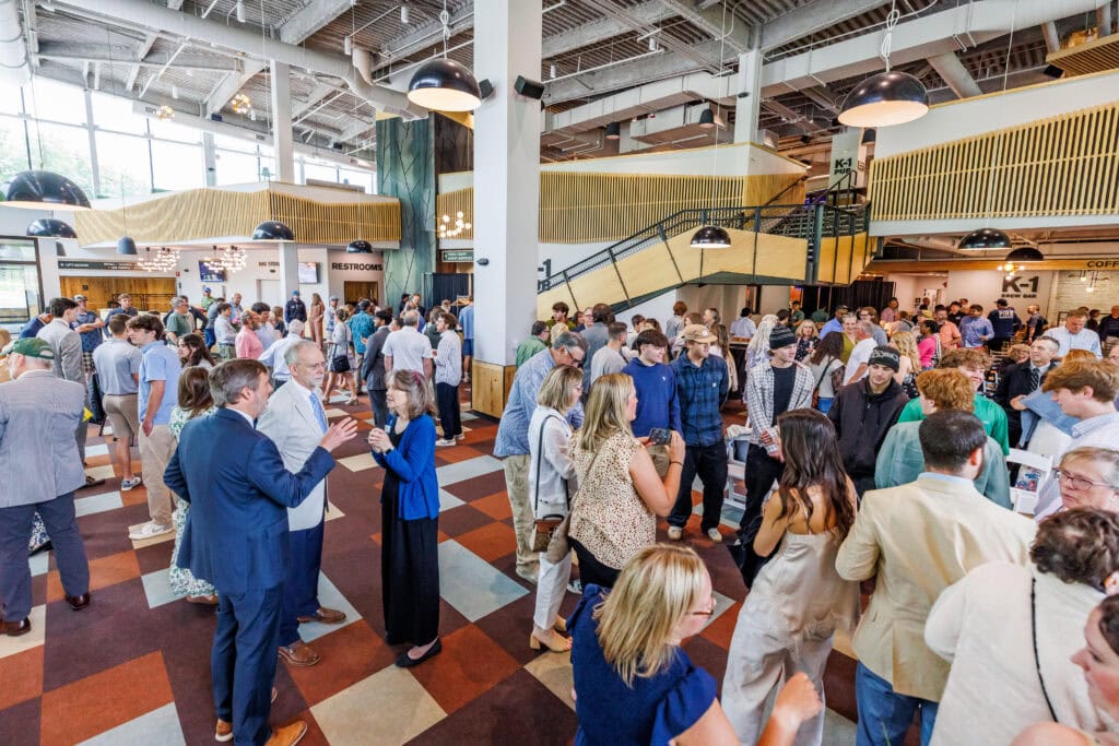 Crowd of people mingling and chatting in a large open lodge lobby with a checkered brown, tan, and cream floor. A metal staircase leads to an upper level with signage for 'K-1 Pub' and 'K-1 Brew Bar' visible in the background.