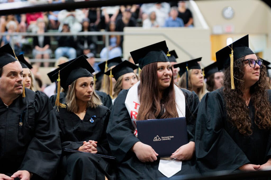 Row of seated graduates in black caps and gowns, viewed from the side. A woman in the center wears a white stole with red lettering and holds a navy folder embossed with the Vermont State University logo. Blurred audience members fill the bleachers behind them.