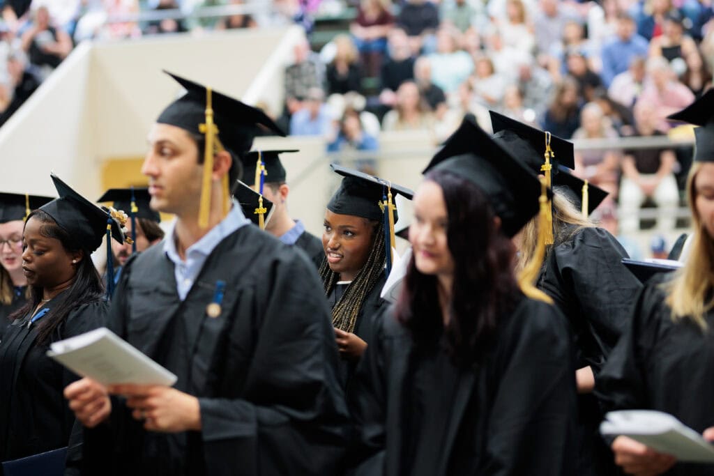 Graduates in black caps and gowns with gold tassels stand in a procession line, some holding programs, with a blurred crowd of seated spectators in the background. A woman with braided hair and a nose ring smiles mid-conversation near the center of the frame.