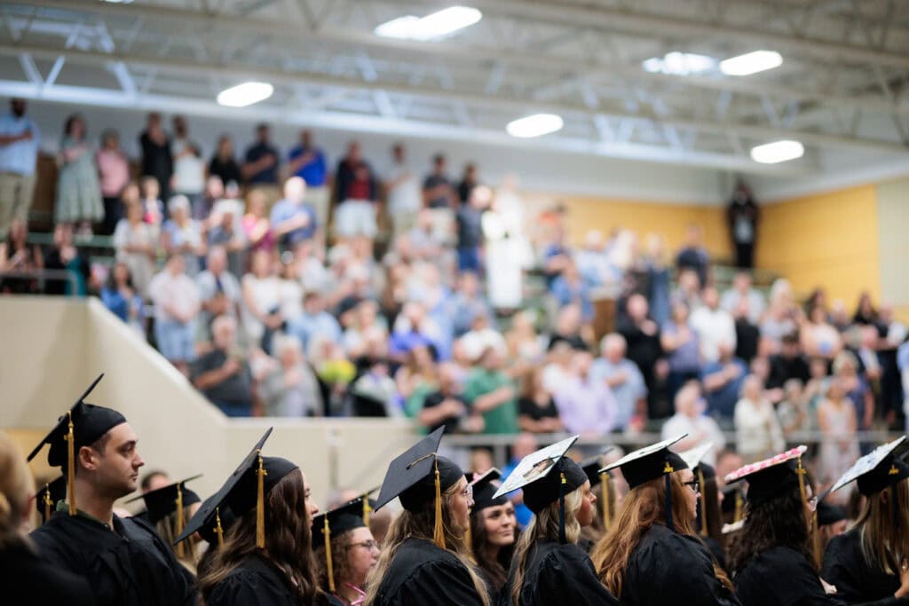 Close-up of graduates in black caps and gowns with gold tassels, seen from behind, facing a packed and blurred audience seated in gymnasium bleachers under fluorescent lighting. Several graduates have decorated the tops of their caps with photos and designs.