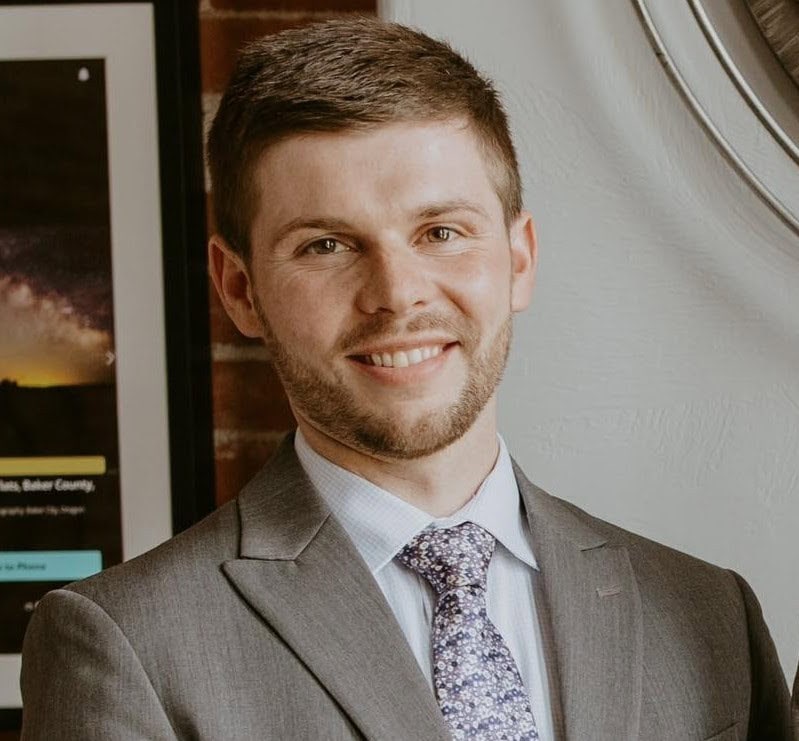 Cody Singer wearing a grey suit and purple tie, smiling at the camera