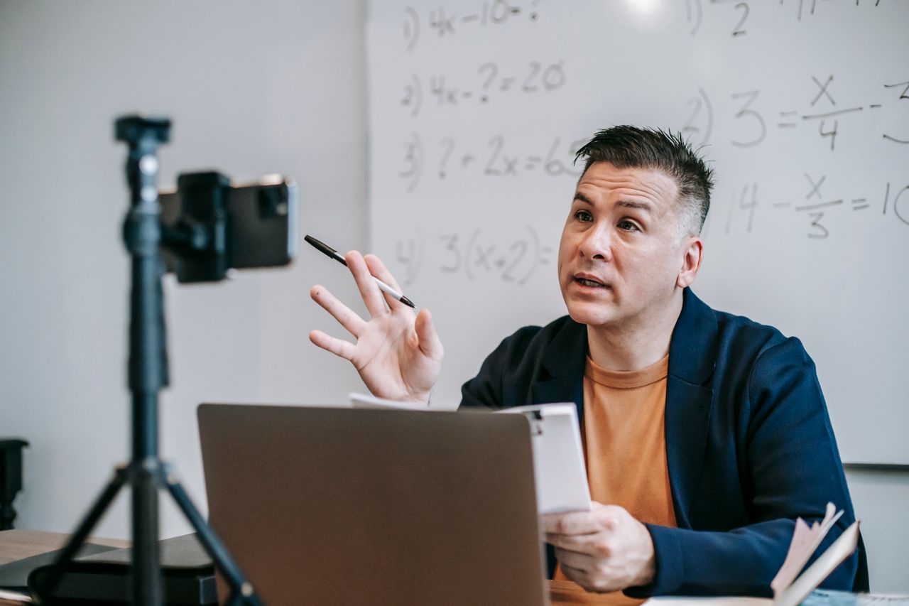 A man sitting at a desk in a classroom. On the desk is a laptop and a phone being held up as a triopod that's being used as a camera. There are math equations on the whiteboard behind him.