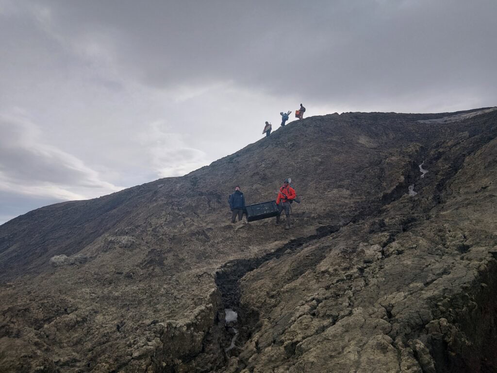 Several researchers traverse a steep, rugged hillside of exposed rock under a dramatic, cloud-filled sky. Two people in the foreground carry a large black equipment case down the slope, while others are visible higher on the ridge carrying field gear. Deep cracks and textured rock formations dominate the barren landscape.