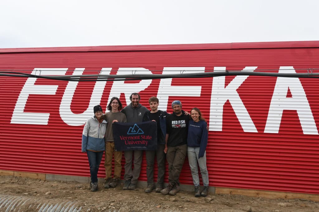 Six field researchers stand side by side in front of a large red corrugated building painted with the word “EUREKA” in oversized white letters. The group holds a Vermont State University banner and wears outdoor clothing and boots. The ground is bare and rocky, and the sky is cloudy.