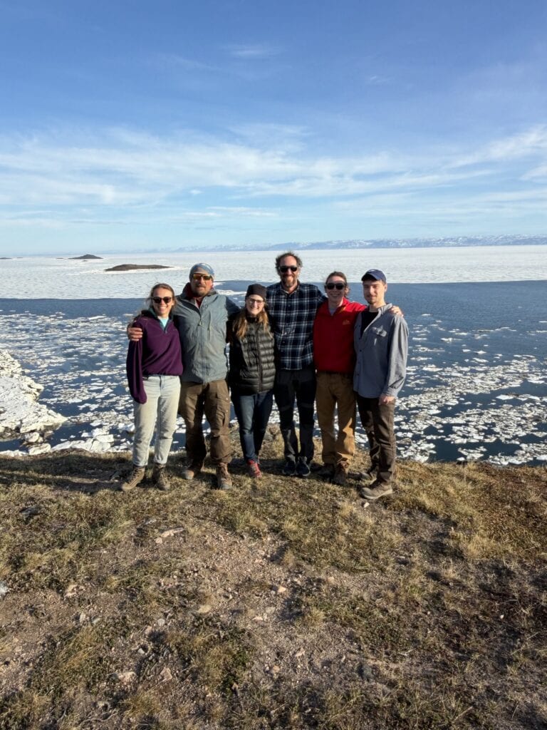 Six people in outdoor gear stand arm in arm on a grassy tundra bluff, smiling at the camera. Behind them, dark open water is scattered with broken ice floes, and snow-dusted mountains line the far horizon under a bright blue sky with thin clouds.