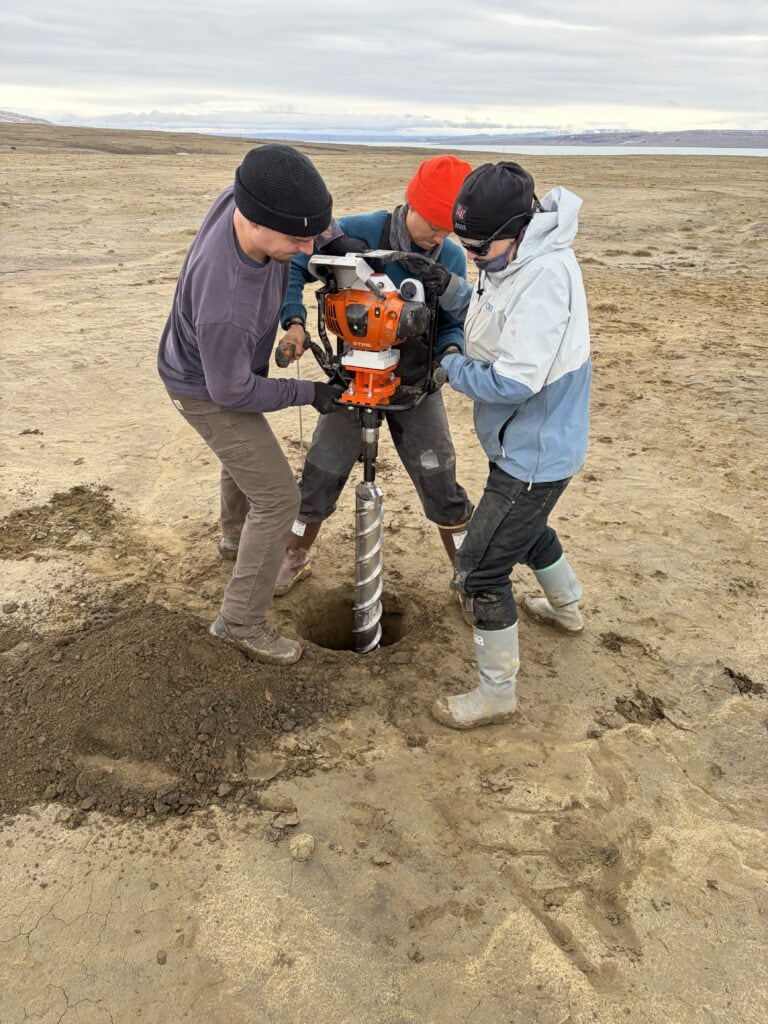 Three people in beanies and rubber boots crowd around an orange gas-powered auger, drilling a hole into bare, sandy tundra soil. A pile of dark excavated soil sits beside the hole, and a flat, barren landscape stretches toward distant water and low hills under an overcast sky.