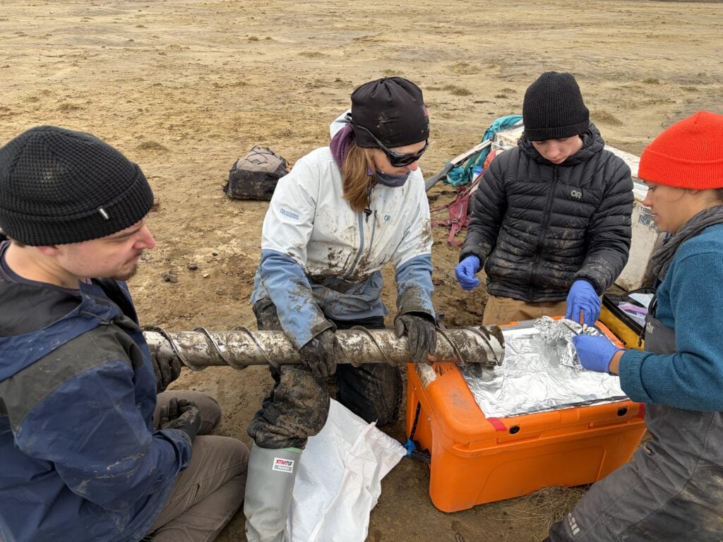 Four people in muddy field clothing kneel around a long steel auger bit, its spiral flights caked with soil, laid across an orange cooler lined with aluminum foil. Two of them wear blue nitrile gloves and handle foil-wrapped samples, while backpacks and other gear sit on the bare ground behind them.