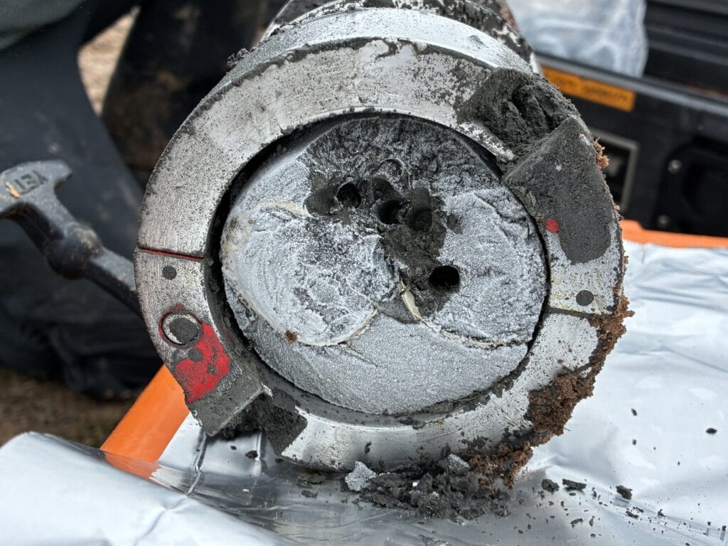 Close-up view of the end of a metal coring tube containing a cylindrical soil and sediment sample. Layers of dark gray, waterlogged material are visible inside the core, with small holes and cracks exposed in the cross section. Mud and soil cling to the outside of the metal tube, which rests on a foil-covered work surface beside field equipment.