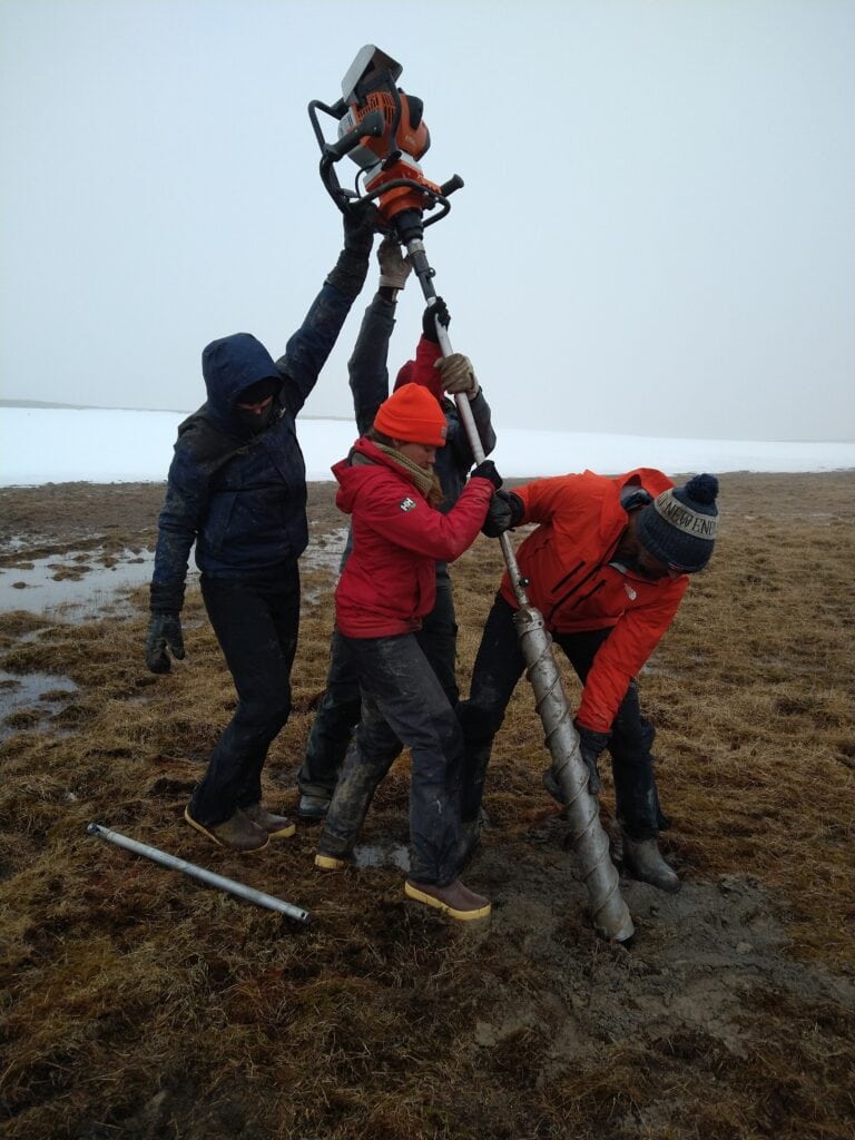Four field researchers work together to operate a large motorized soil auger in a wet Arctic tundra environment. Wearing cold-weather gear, boots, and gloves, the team steadies the drill as it bores into thawed ground near patches of lingering snow. The landscape is flat and treeless, with a gray, foggy sky stretching across the background.