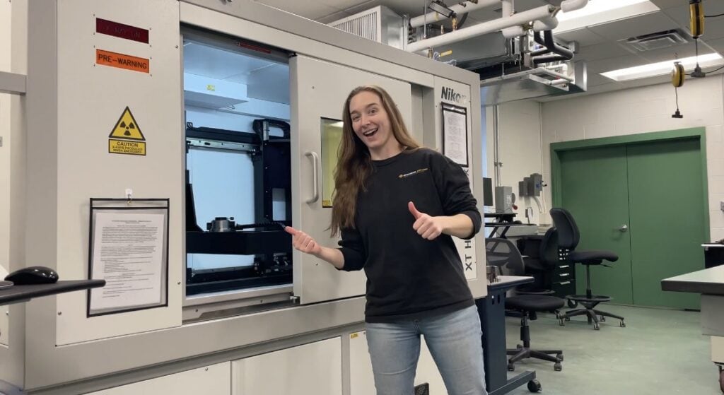 Person stands beside a large industrial inspection or manufacturing machine in a laboratory setting, giving a thumbs-up gesture. The machine's access door is open, revealing the interior workspace. Safety signage is mounted on the machine, and computer workstations, office chairs, exposed piping, and green double doors are visible throughout the room. The image highlights hands-on work with advanced manufacturing and quality control equipment in a technical learning environment.