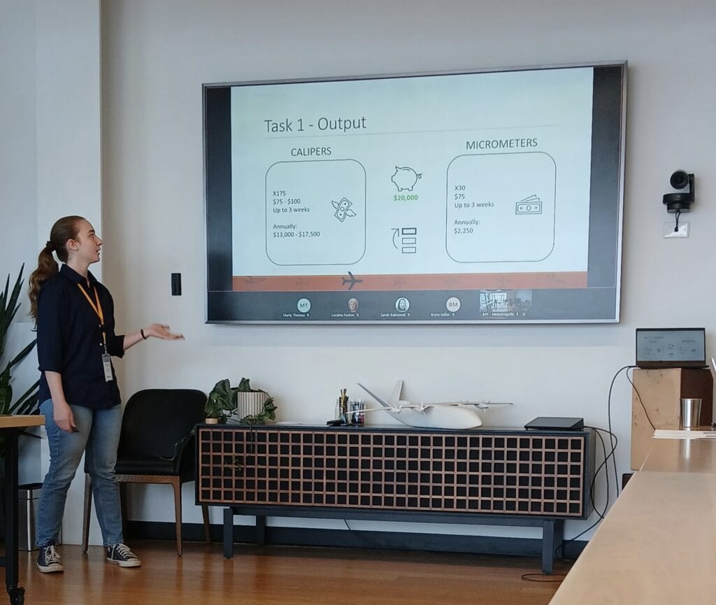 Person giving a presentation in a conference or classroom setting while standing beside a large display screen. The slide shown is titled “Task 1 - Output” and includes sections labeled “CALIPERS” and “MICROMETERS” with cost and quantity information. A model aircraft sits on a cabinet beneath the screen, and a laptop is positioned on a nearby table connected to the presentation display.