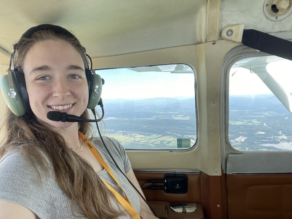 Person wearing an aviation headset with microphone sits inside a small aircraft cabin. Through the aircraft windows, a broad landscape of fields, forests, and distant mountains is visible below. The interior includes beige and brown paneling, a seatbelt, and aircraft windows framing the aerial view.
