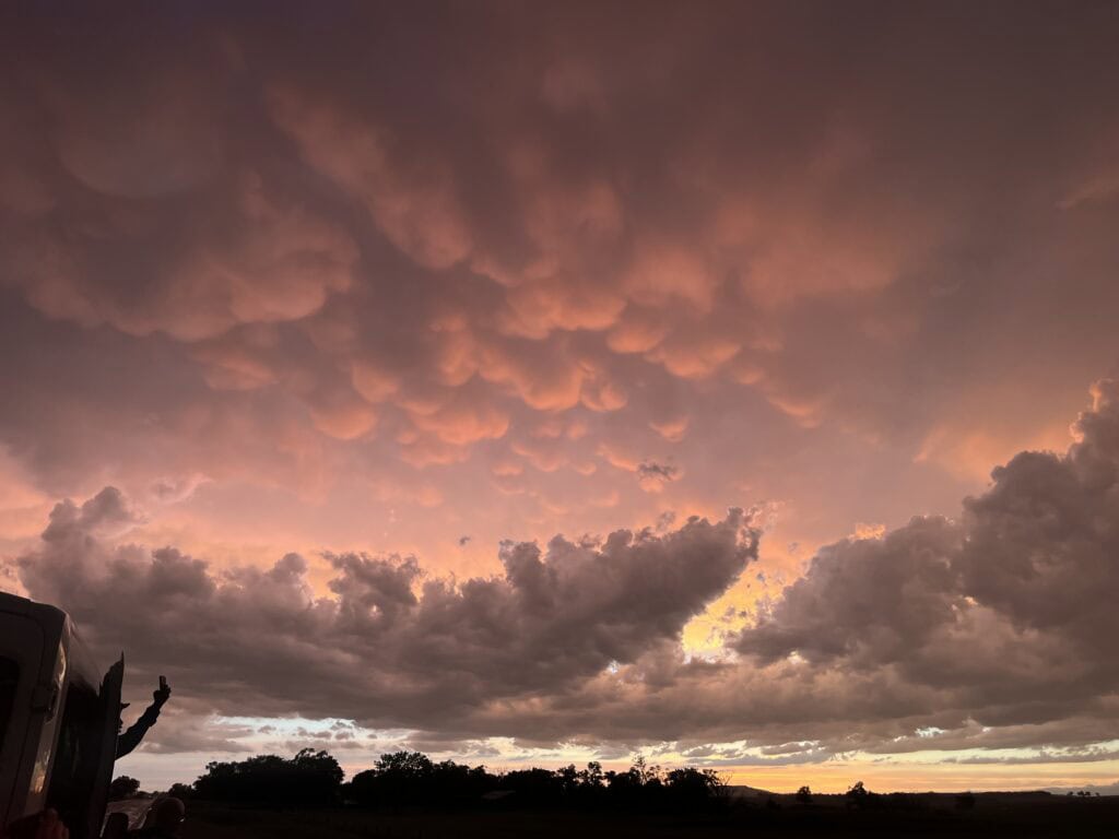 Alt text: Dramatic sunset sky filled with pink and orange mammatus clouds above a dark rural landscape. Silhouetted storm observers stand beside a vehicle at the left edge of the image, watching the illuminated cloud formations near the horizon.