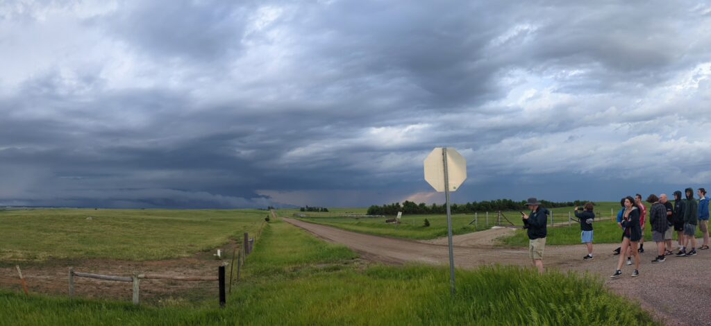 Alt text: Wide view of a severe storm over open grassland and farmland. Dark layered storm clouds stretch across the sky above a rural dirt-road intersection, where a group of storm observers stands along the roadside watching the approaching weather.