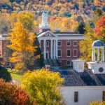 A scenic fall view of the Vermont State University Castleton campus, featuring a red-brick academic building with white columns and a central bell tower surrounded by vibrant autumn foliage in shades of gold, orange, red, and green. In the foreground, a white historic building with a domed cupola and clock is partially framed by colorful trees, with forested hills covered in peak fall colors rising in the background.